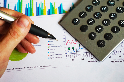 A printed page with graphs and charts partially covered by a calculator, a hand with a pen is poised to write on the page