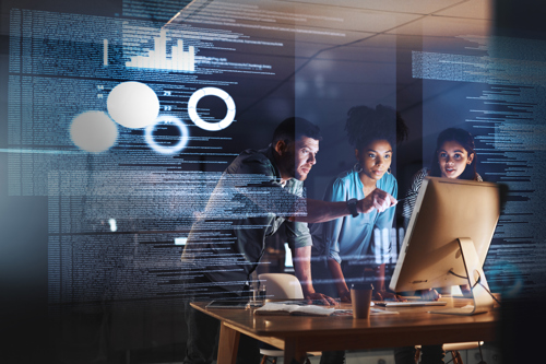 Three students collaborate around a computer screen in an office, one of them is pointing at the screen