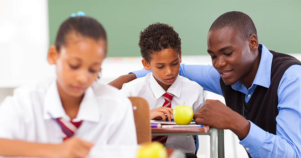 Two students in a classroom with the teacher speaking to one of them, helping them solve a problem